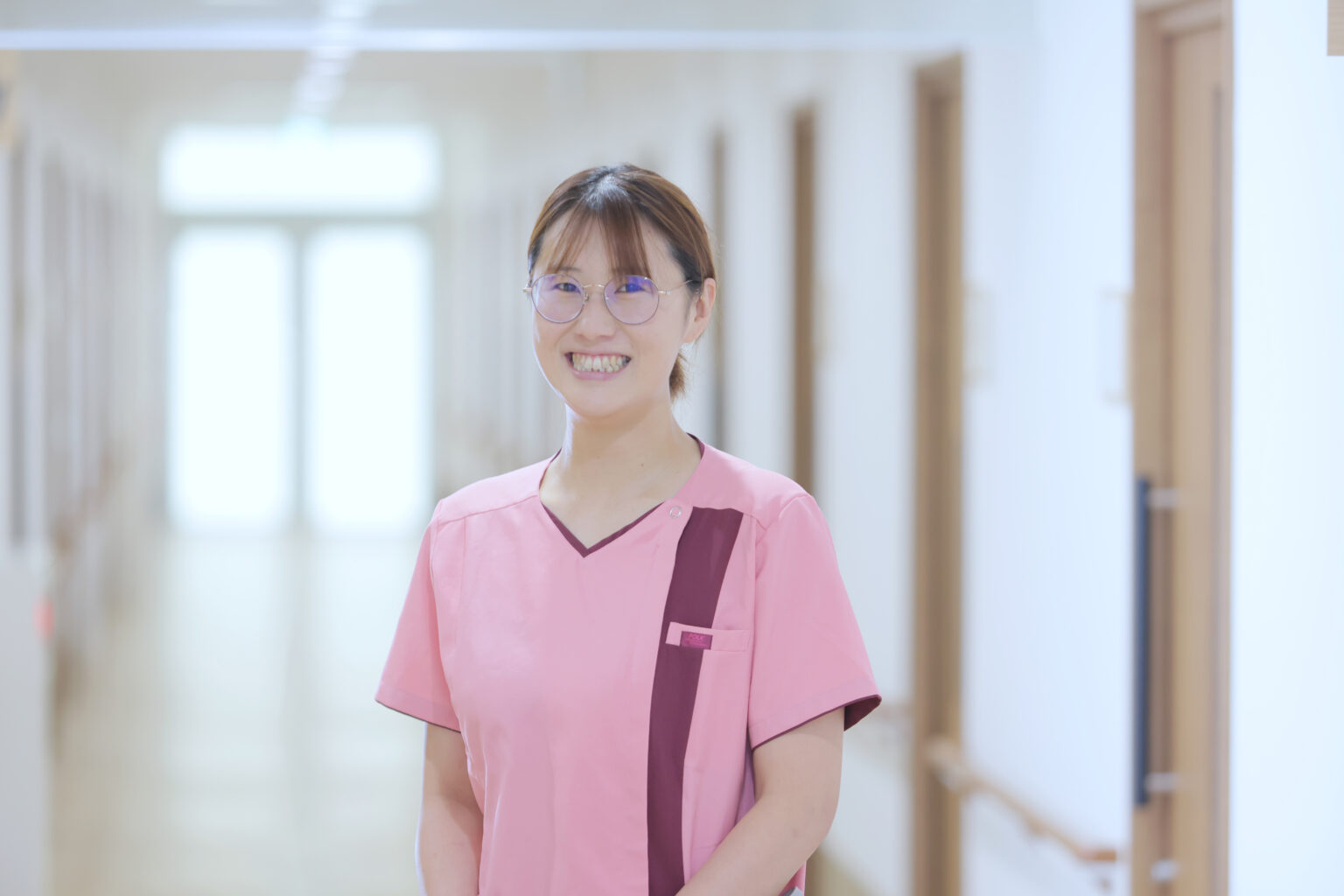 介護施設の廊下に立つ笑顔のスタッフ。ピンク色のユニフォーム姿。
